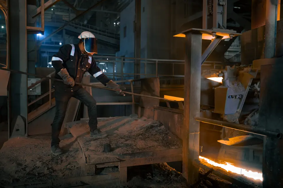 A worker in a heat-proof jacket and face shield works with a long rod beside glowing metal