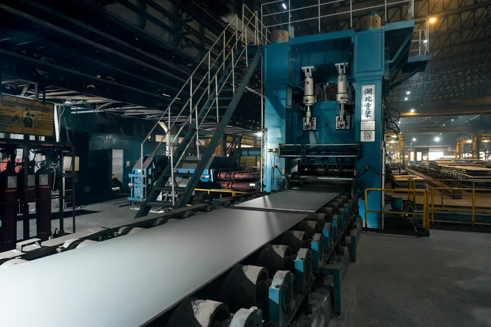 A steel plate on a line of rollers in a large plant, with stairs and walkways above