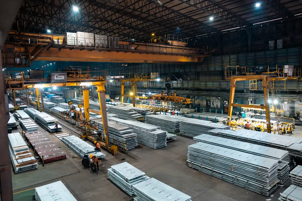 Stacks of steel plates and overhead cranes in a huge factory bay, seen from above