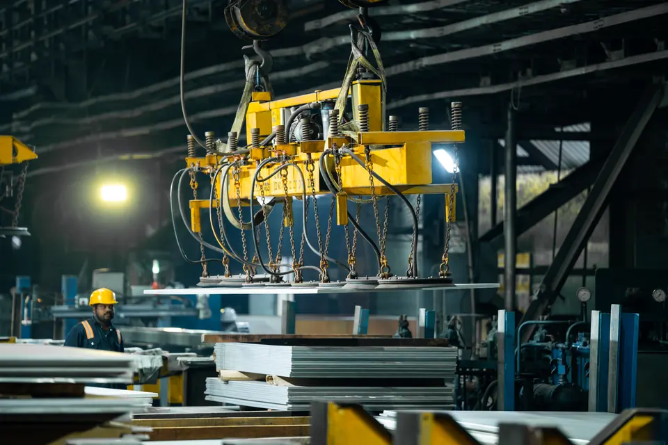 A yellow lifter carries a steel plate while a worker in a hard hat watches
