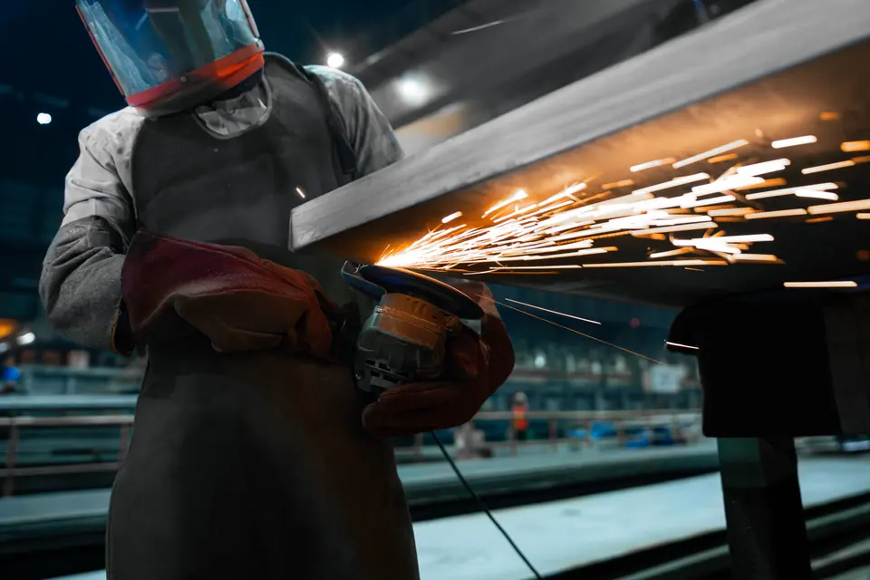A worker in a face shield grinds the edge of a steel plate, sending sparks flying
