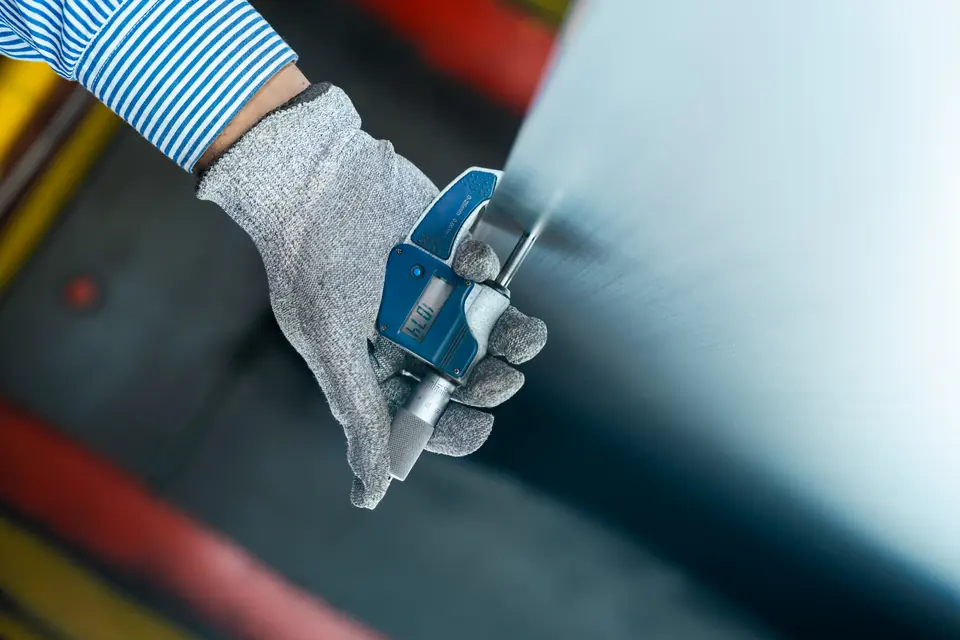 A gloved hand measures a steel sheet with a micrometer