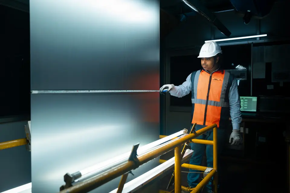 An inspector in a hard hat measures a large, bright steel sheet with a tape