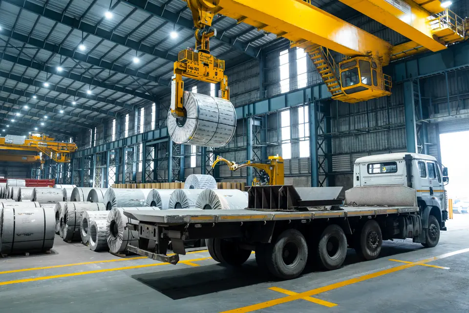 A crane lowers a wrapped steel coil onto a truck, with rows of coils beside it