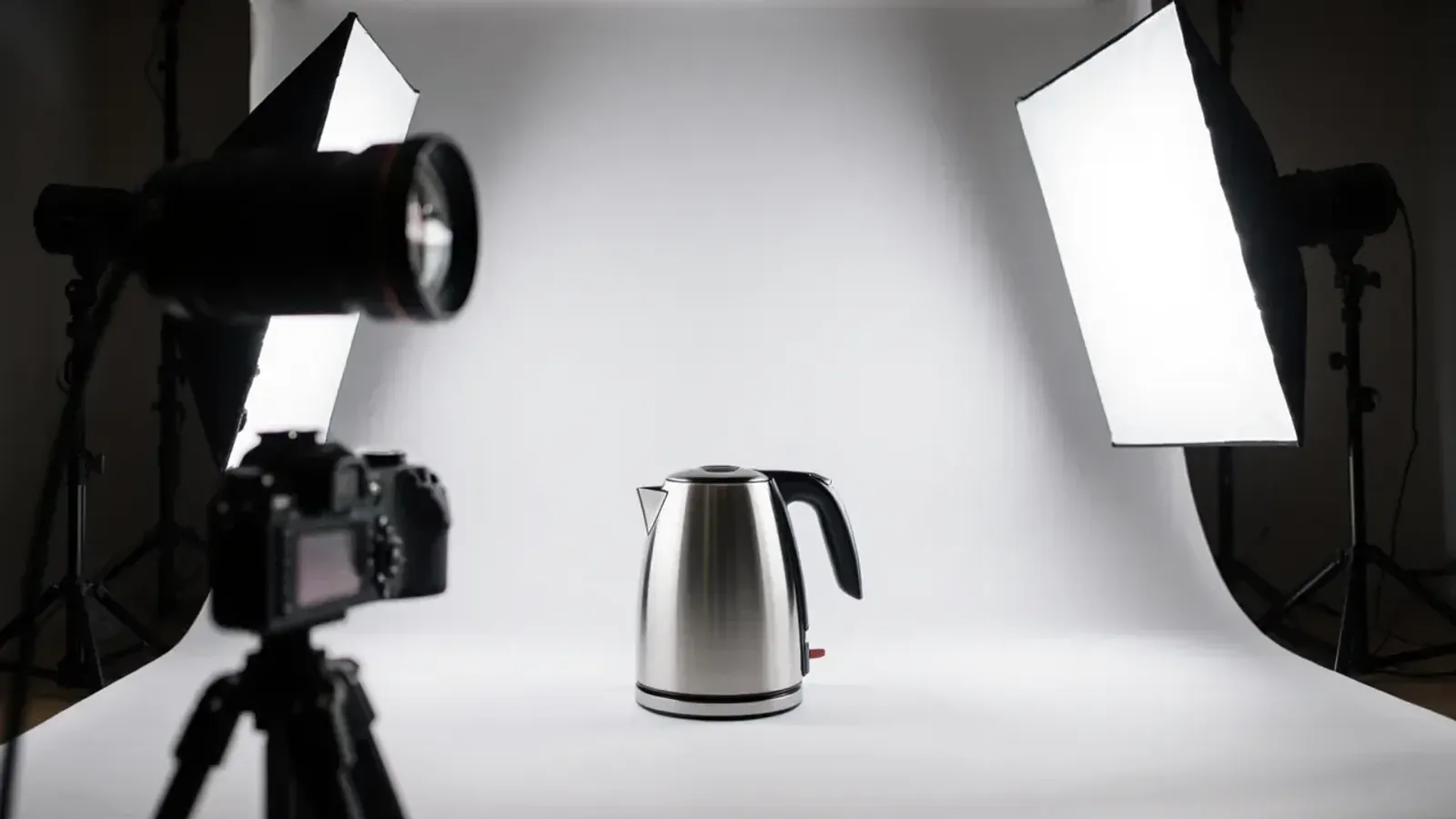 A steel electric kettle on a white paper backdrop between two softbox lights, with a camera on a tripod in the foreground