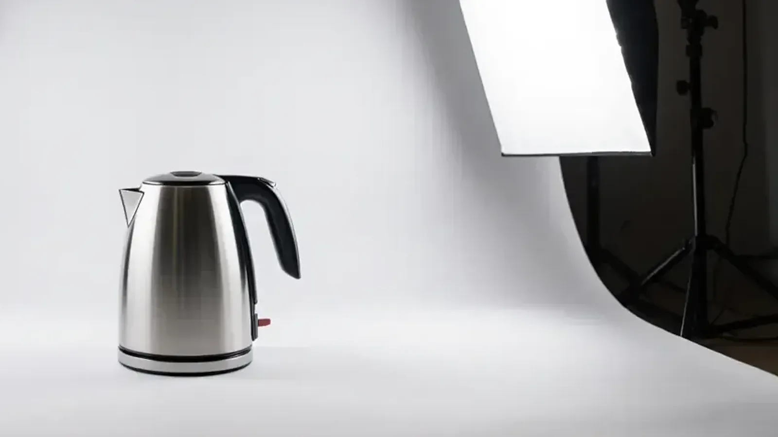 A steel electric kettle on a white paper backdrop, lit by a softbox on a stand