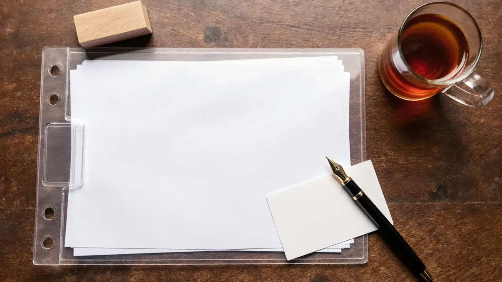 Overhead view of blank papers in a clear folder, a blank card, a fountain pen, a wooden stamp and a glass of tea on a wooden desk