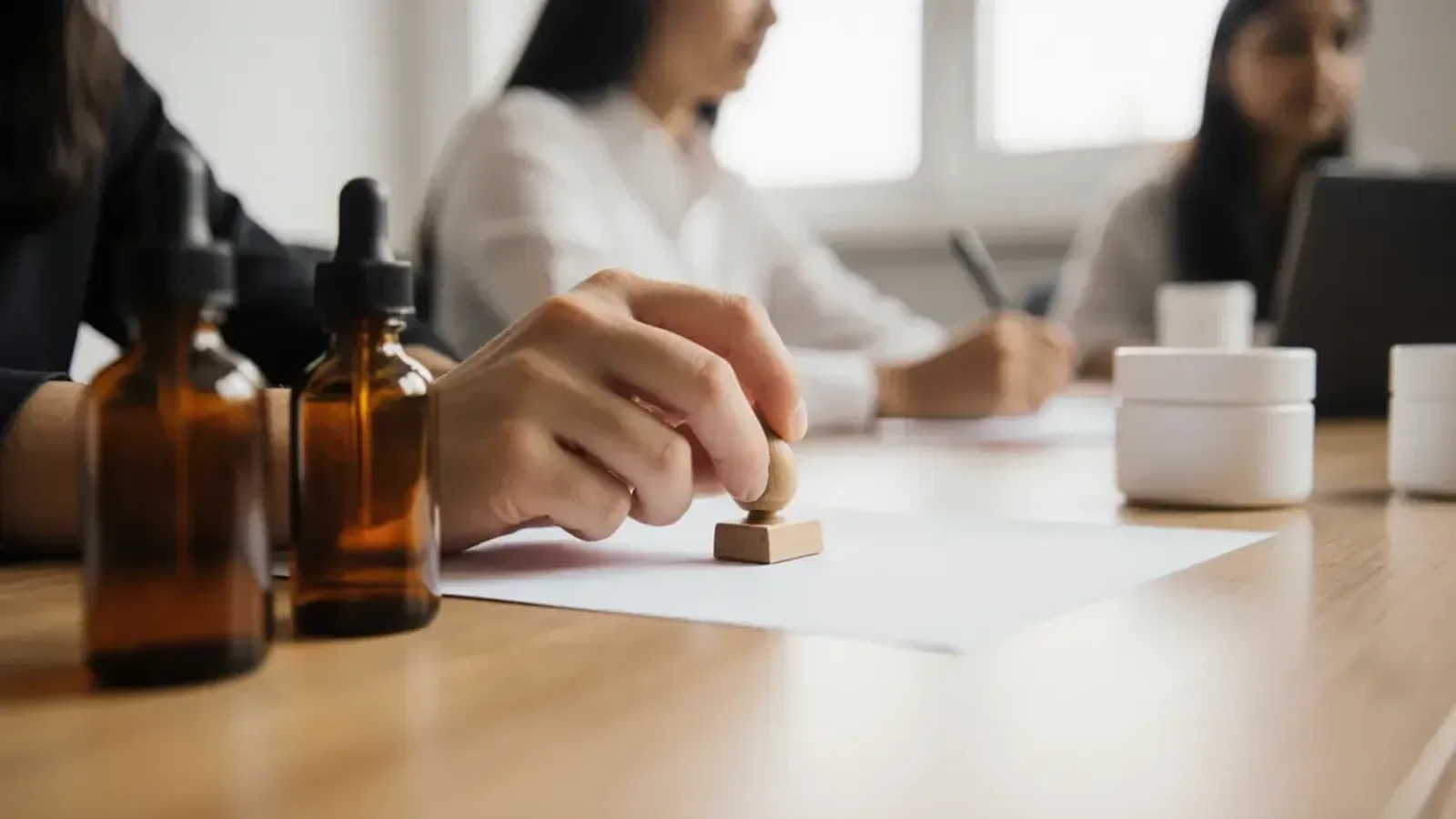 A hand pressing a wooden rubber stamp onto a sheet of paper on an office desk, beside amber glass dropper bottles and white jars, with colleagues working behind