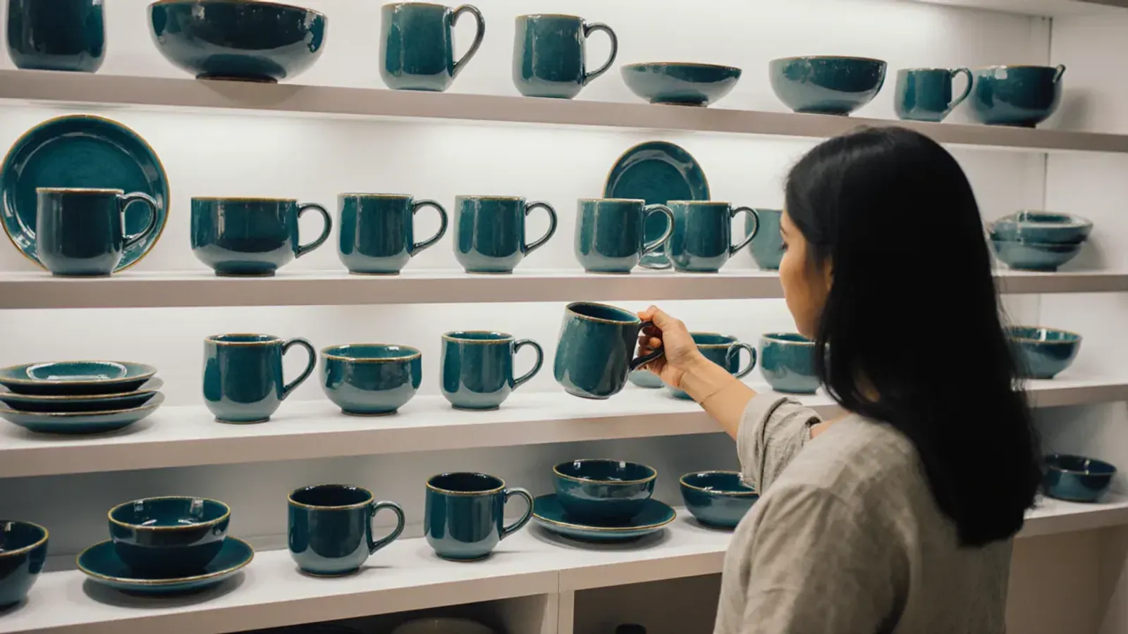 A young woman placing a teal mug on white shelves lined with a ceramics brand's full range of matching mugs, bowls and plates