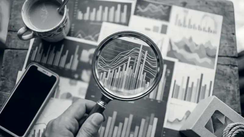 A hand holding a magnifying glass over printed sales charts, beside a cup of chai, a phone and a product box