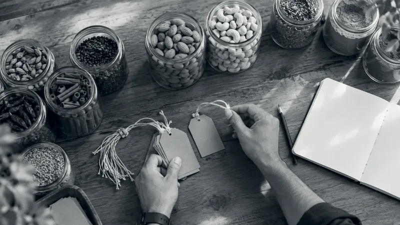 Hands tie blank kraft price tags with cotton string beside glass jars of cardamom, cinnamon, almonds, cashews and ground spices on a wooden table, with an open notebook and a pencil