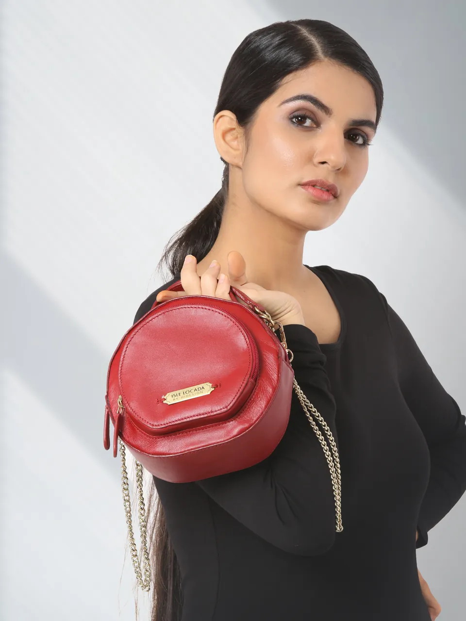 A model in a black top holding up a round red bag on a gold chain, in soft window light