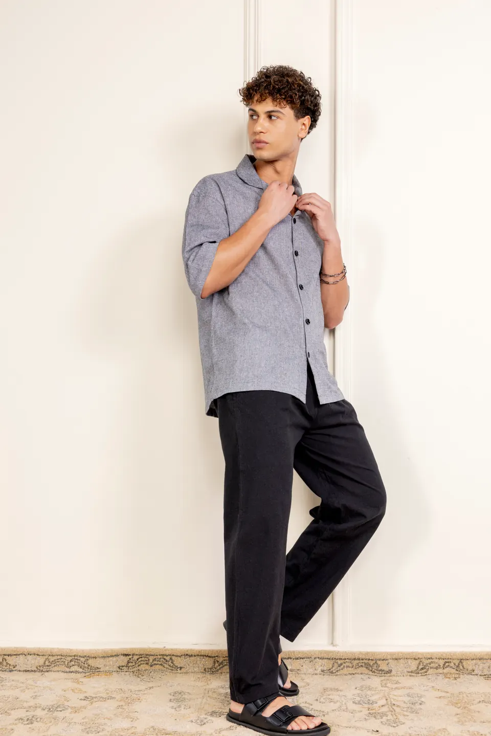 A male model in a grey linen shirt and black trousers, leaning against a cream wall