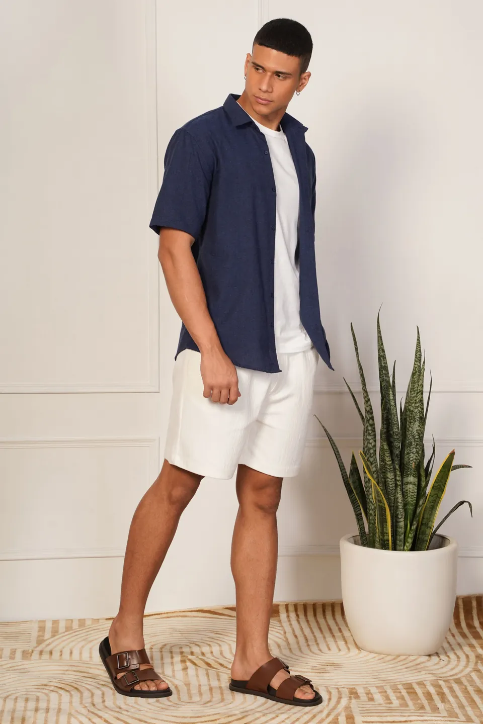 A male model in a navy shirt and white shorts beside a plant, in a styled room