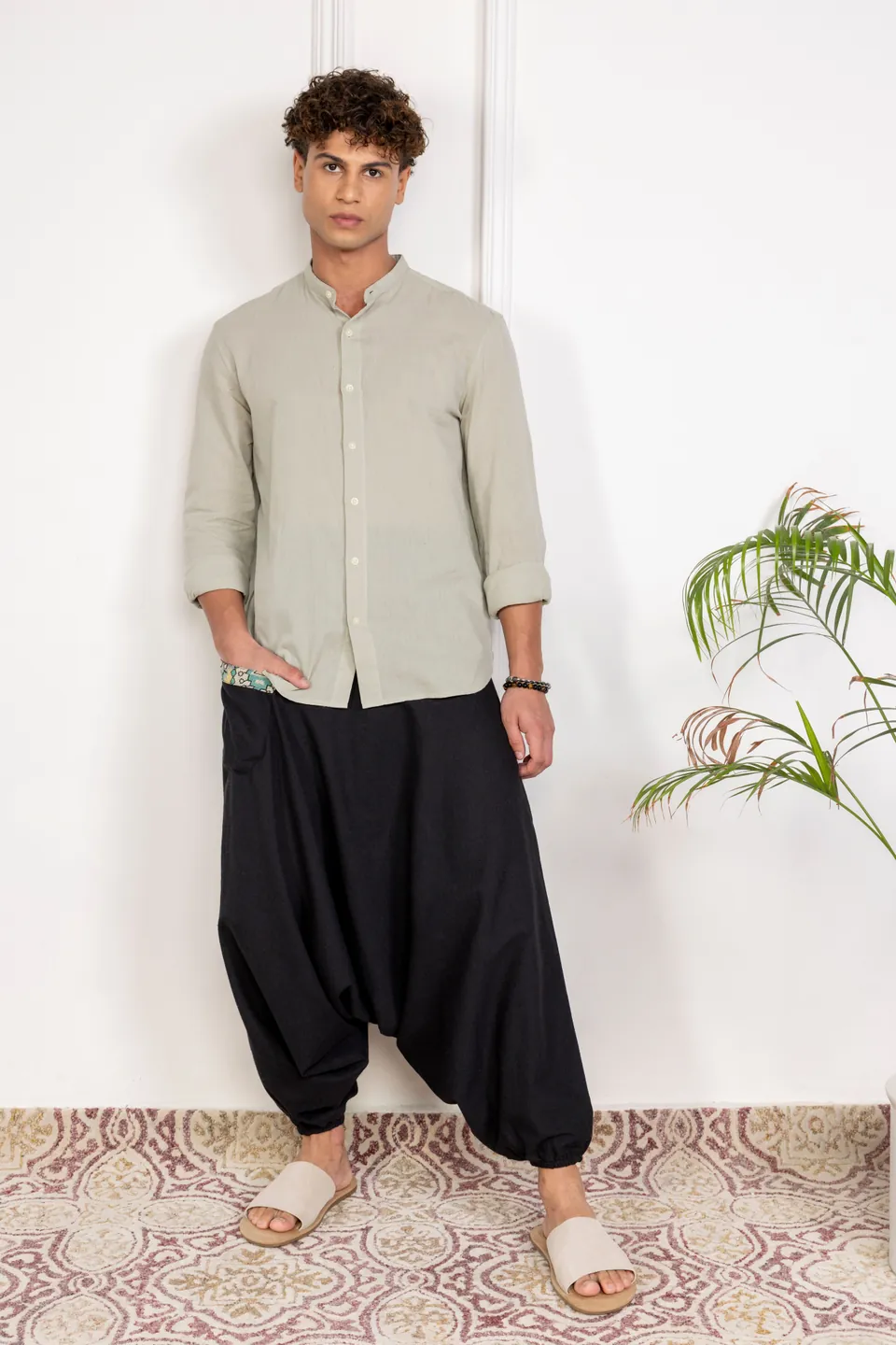 A male model in a sage shirt and black harem trousers, standing beside a plant