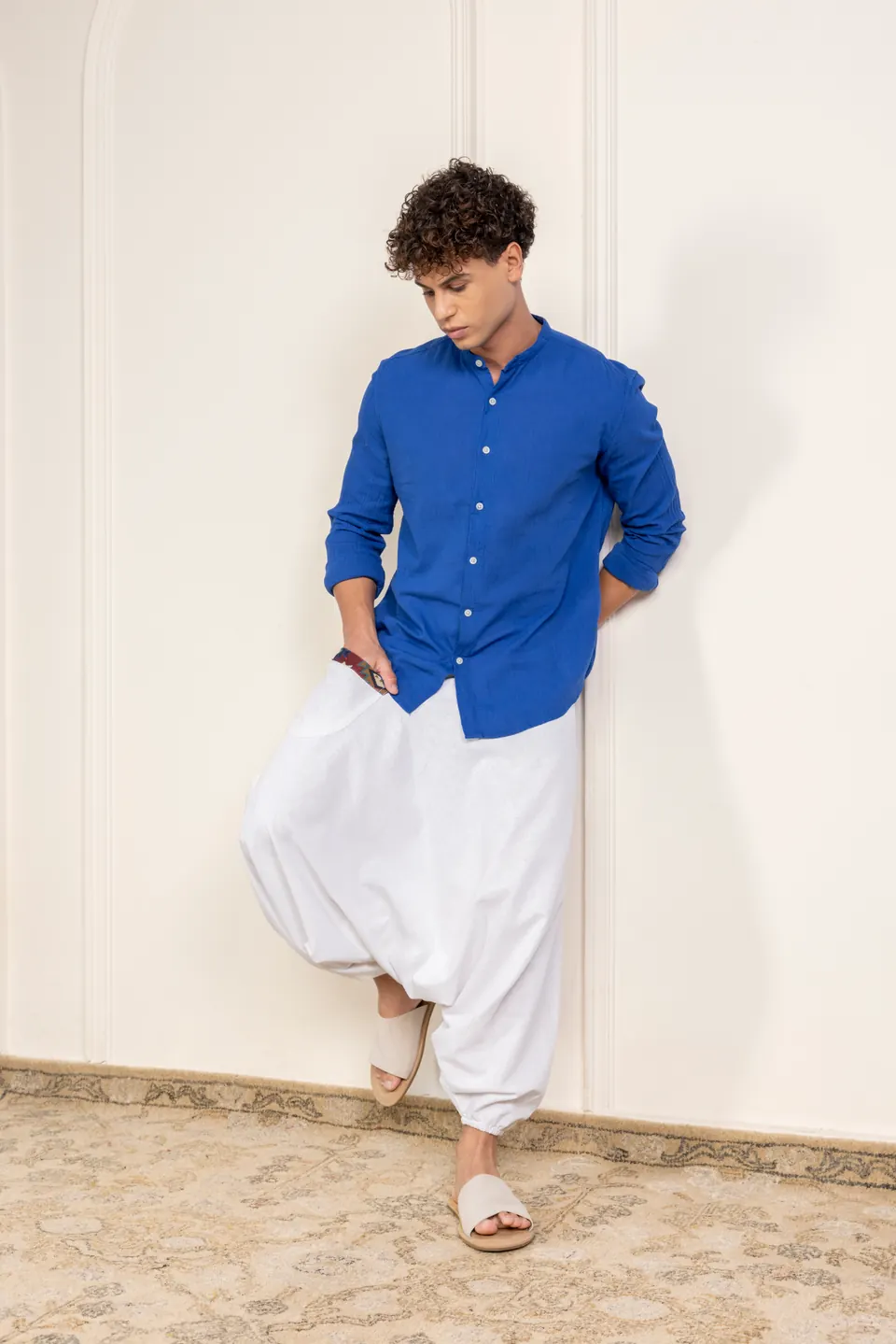 A male model in a blue shirt and white harem trousers, leaning against a wall