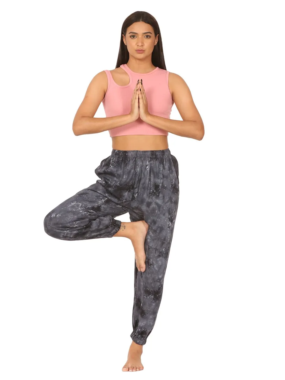 A model in a pink crop top and grey tie-dye harem pants, standing in a yoga tree pose with her hands together, on white