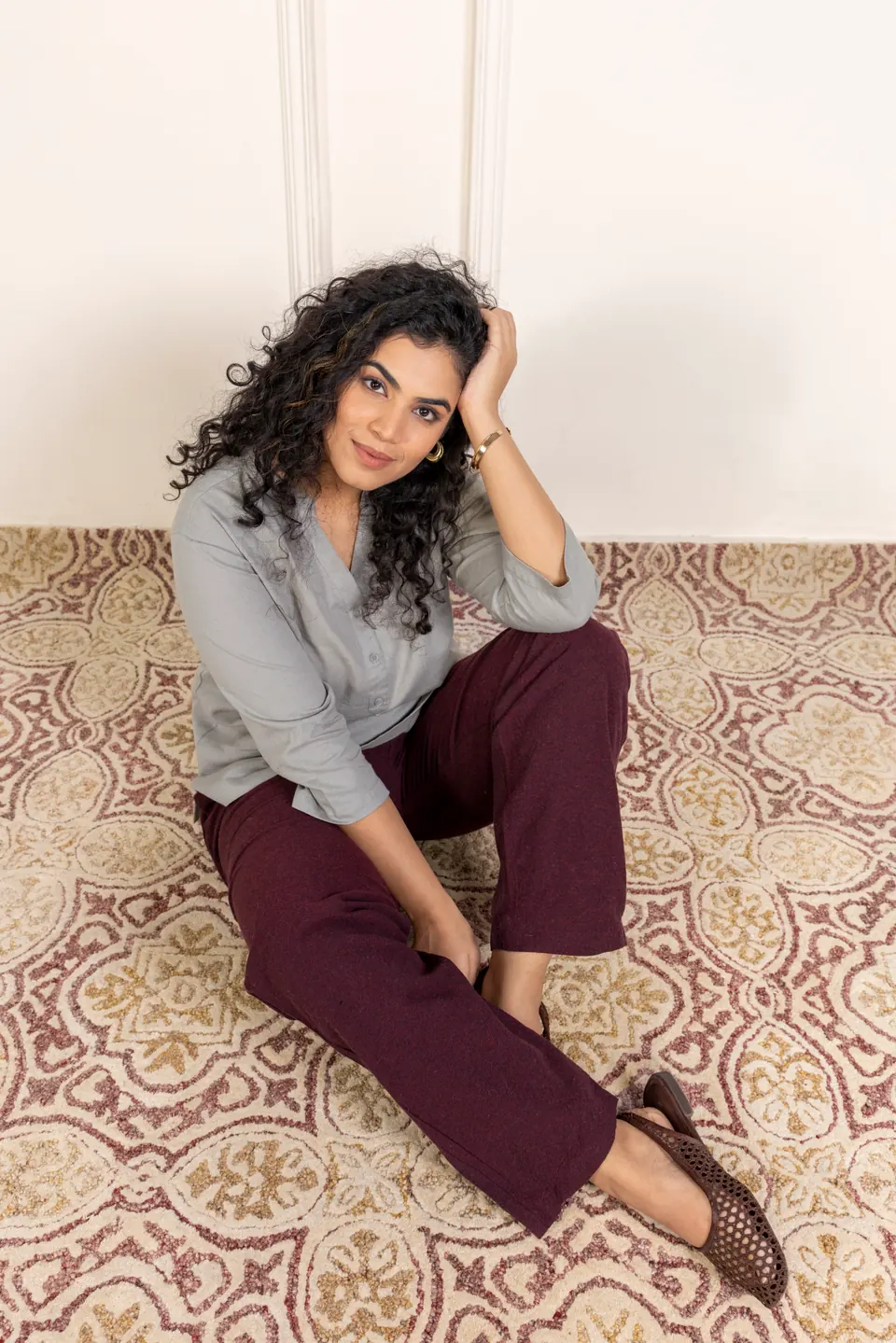 A female model in a grey shirt and plum trousers, seated on a patterned rug