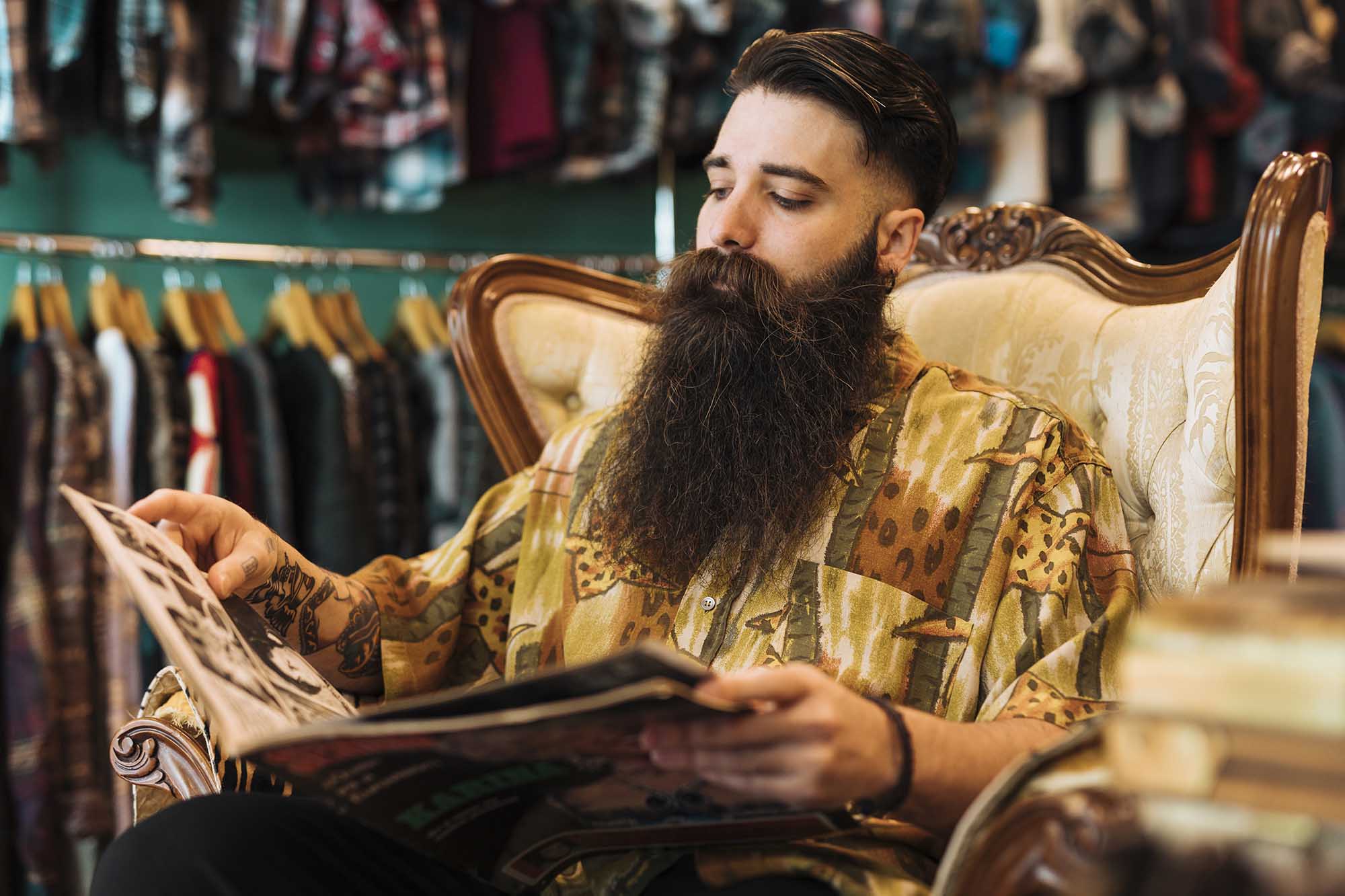 bearded-fashionable-young-man-sitting-chair-looking-magazine-shop